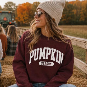 May include: A maroon crewneck sweatshirt featuring the white text PUMPKIN SEASON in a collegiate-style font across the chest. The garment is paired with a beige ribbed knit beanie and dark-tinted sunglasses, creating a casual look.
