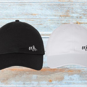 K&ouml;nnte beinhalten: Eine schwarze Baseballkappe mit dem Wort &bdquo;Mr.&ldquo; in wei&szlig;er Schrift neben einer wei&szlig;en Baseballkappe mit dem Wort &bdquo;Mrs.&ldquo; in wei&szlig;er Schrift. Die Kappen befinden sich auf einer hellblauen und wei&szlig;en Holzoberfl&auml;che.