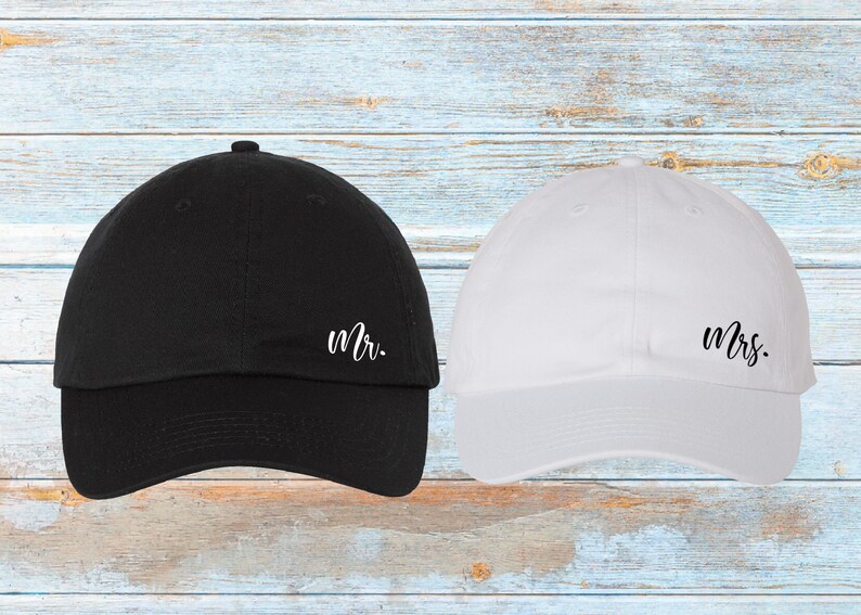 K&ouml;nnte beinhalten: Eine schwarze Baseballkappe mit dem Wort &bdquo;Mr.&ldquo; in wei&szlig;er Schrift neben einer wei&szlig;en Baseballkappe mit dem Wort &bdquo;Mrs.&ldquo; in wei&szlig;er Schrift. Die Kappen befinden sich auf einer hellblauen und wei&szlig;en Holzoberfl&auml;che.