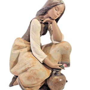 May include: A ceramic figurine of a woman kneeling, resting her chin on her hand. She wears a long-sleeved cream-colored shirt, a brown vest, and a long skirt. She holds a small pink rose and is next to a brown jug.