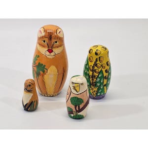 May include: A set of four wooden nesting dolls, each painted with a different animal design. The largest doll features a cat, while the others depict an owl, an eagle, and a squirrel. The dolls are painted in shades of brown, orange, yellow, and green.