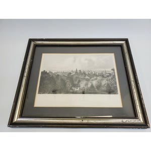 May include: Black and silver framed print of a city landscape. The black and white artwork depicts a detailed view of a city with buildings and trees. The frame has a silver inner edge and a black outer edge.