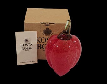 May include: A red glass pepper sculpture with a green stem, next to a cardboard box and a white card with the text "KOSTA BODA". The pepper is a vibrant red color and has a glossy finish. The sculpture is likely a decorative item.