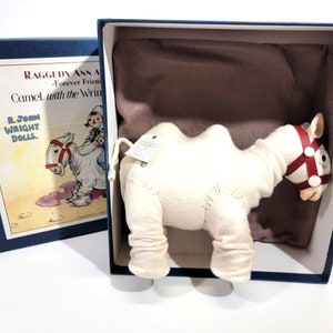 May include: A white plush camel with a red leather bridle, sitting in a blue box with a Raggedy Ann and Andy illustration on the side. The illustration shows Raggedy Ann riding the camel. The box reads "Raggedy Ann and Andy - Forever Friends - Camel with the Wrinkles".