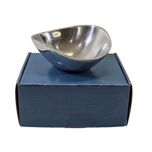 May include: A small, polished silver bowl with a unique, asymmetrical shape. The bowl is set on a dark blue cardboard box. The bowl's interior reflects light, highlighting its smooth surface. The overall presentation suggests a decorative or serving piece.