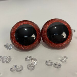 Puede incluir: Dos ojos saltones grandes, redondos y de color rojo con purpurina, con pupilas negras. Los ojos est&aacute;n sobre una superficie blanca con peque&ntilde;os cristales transparentes en forma de diamante esparcidos a su alrededor.