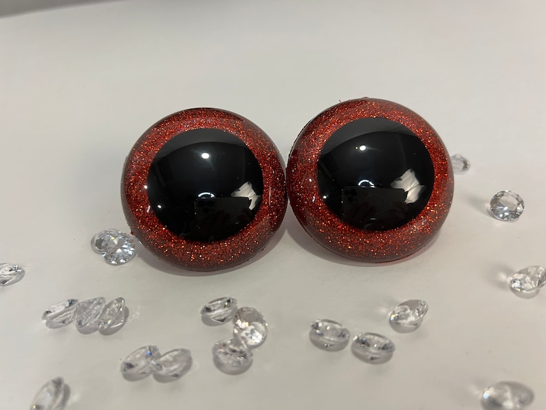 Puede incluir: Dos ojos saltones grandes, redondos y de color rojo con purpurina, con pupilas negras. Los ojos est&aacute;n sobre una superficie blanca con peque&ntilde;os cristales transparentes en forma de diamante esparcidos a su alrededor.
