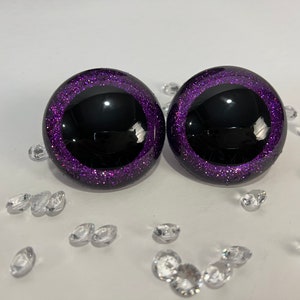 Puede incluir: Dos ojos de pl&aacute;stico negro con un borde de purpurina morado. Los ojos son redondos y tienen una pupila negra.