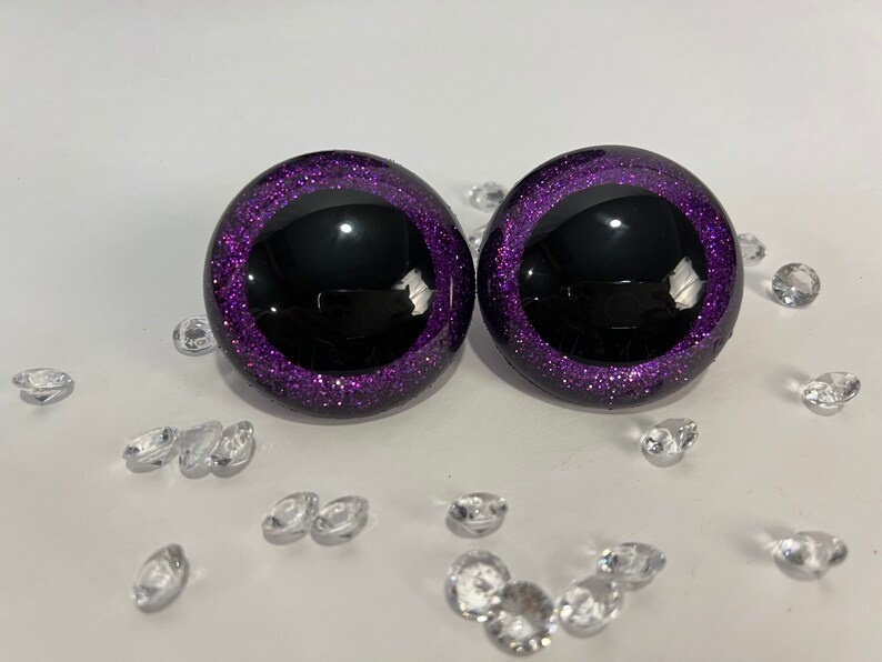 Puede incluir: Dos ojos de pl&aacute;stico negro con un borde de purpurina morado. Los ojos son redondos y tienen una pupila negra.
