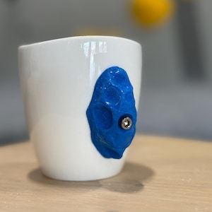 Peut inclure: Mug en céramique blanche avec une prise d'escalade bleue texturée fixée. La prise d'escalade a un boulon au centre. Le mug est posé sur une surface en bois brun clair.