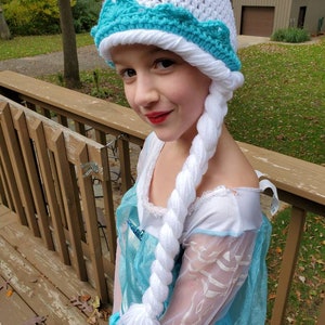 May include: A white crocheted hat with a blue crocheted crown and a long white braid attached. The hat is worn by a child dressed as Elsa from the movie Frozen.