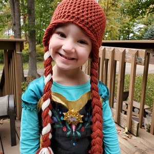 May include: A young girl wearing a red crocheted hat with long braids, dressed in a blue and black dress with a snowflake design.