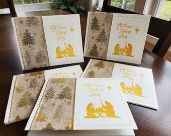 May include: Five white Christmas cards with gold and brown patterned ribbon. Each card features a gold silhouette of the nativity scene and the text "Oh Come Let Us Adore Him".