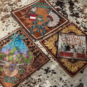 May include: Three square, hand-tooled leather pieces with unique designs. One features a rodeo scene, another an American Indian headdress, and the third a hand with scissors and cacti. Each piece has personalized text.