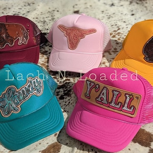 May include: Five trucker hats with different designs. The hats are in various colors, including pink, teal, maroon, yellow, and hot pink. The hats have different embroidered patches, including a buffalo, a steer head, a spade, the word "Howdy", and the words "Y'all".