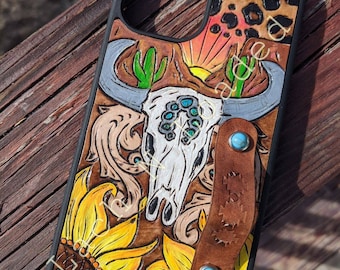 May include: A leather phone case with a western design. The case features a skull, sunflowers, cacti, and a leopard print. The case has a brown leather strap with turquoise accents.