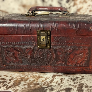 Könnte beinhalten: Eine Vintage-Holzkiste aus braunem Leder mit einem geprägten Blumenmuster und einem Messingverschluss und -griff.