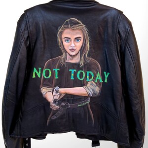 May include: Black leather jacket with a painted portrait on the back. The portrait features a woman with blue eyes and the words "NOT TODAY" in green. The jacket has a zipper closure and a belt detail.