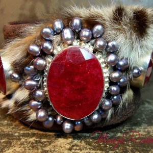 May include: A silver bracelet with a large red gemstone surrounded by a ring of smaller purple gemstones. The bracelet is adorned with faux fur.