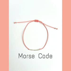 Puede incluir: Un brazalete ajustable de cuerda de color coral con cuentas plateadas que deletrean "Morse Code" en código Morse.