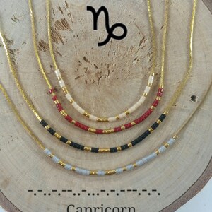Puede incluir: Collar de cadena dorada con cuatro capas de cuentas en rojo, blanco, negro y gris. El collar presenta un símbolo del zodíaco Capricornio y la palabra "Capricornio" escrita en código Morse.