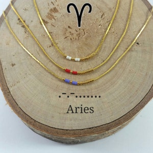 Puede incluir: Collar de cadena dorada con un pequeño dije de cuentas con cuentas azules, rojas y blancas. El dije tiene la forma del símbolo astrológico de Aries, que son cuernos de carnero. El collar se muestra sobre una superficie de madera con la palabra "Aries" y el símbolo braille de Aries.