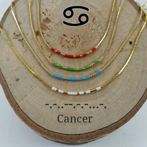 Puede incluir: Collar de cadena dorada con cuatro hebras de cuentas de diferentes colores. Las cuentas son rojas, verdes, azules y blancas. El collar se muestra sobre una superficie de madera con el símbolo del zodíaco Cáncer y la palabra "Cáncer" escrita en código Morse.