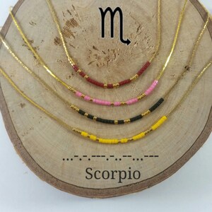 May include: Four gold-tone necklaces with beaded pendants in red, pink, black, and yellow. Each necklace features a curved row of beads with gold accents. The word "Scorpio" is below the necklaces, along with the Scorpio symbol.