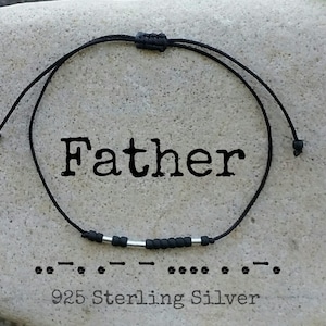 Puede incluir: Pulsera de cordón negro con una barra de plata y cuentas que deletrean "Padre" en braille. La pulsera está sobre un fondo de piedra gris. Plata de ley 925.