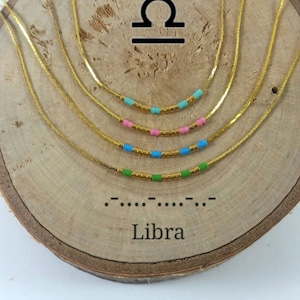 Puede incluir: Cuatro collares de cadena dorada con cuentas de diferentes colores, superpuestos sobre una superficie de madera. Las cuentas son verdes, azules, rosas y turquesas. El símbolo del zodíaco de Libra y la palabra "Libra" están escritos debajo de los collares.