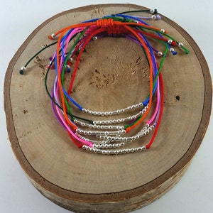 Può includere: Un mucchio di braccialetti dell'amicizia colorati con perline d'argento. I braccialetti sono fatti di cordoncino sottile e sono legati insieme in un nodo.