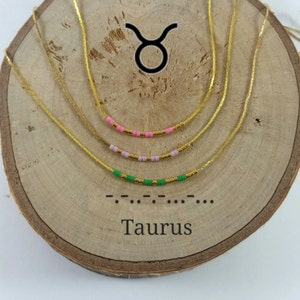 Puede incluir: Un collar de cadena dorada con tres filas de cuentas de colores. Las cuentas son verdes, rosas y doradas. El collar se muestra sobre una superficie de madera con el símbolo del zodíaco Tauro y la palabra "Tauro" escrita debajo.