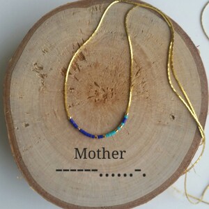 Puede incluir: Un collar de cadena dorada con una pequeña fila de cuentas azules, turquesa y doradas. El collar se muestra sobre una superficie de madera con la palabra "Mother" escrita debajo.