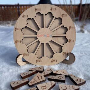 Puede incluir: Un c&iacute;rculo de madera con un dise&ntilde;o de nota musical y 12 ranuras alrededor del borde. Hay 12 piezas de madera con notas musicales y letras en ellas. Las piezas est&aacute;n esparcidas sobre una superficie blanca.