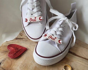 Puede incluir: Un par de zapatos de lona blancos con detalles de corazón rojo y la palabra "Oui" escrita en cada zapato. También se puede ver un corazón rojo y dos anillos de oro en la imagen.