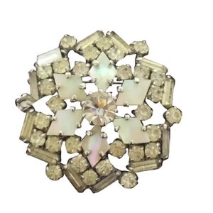 Puede incluir: Un broche de tono plateado con un diseño de copo de nieve. El copo de nieve está hecho de diamantes de imitación transparentes e iridiscentes.
