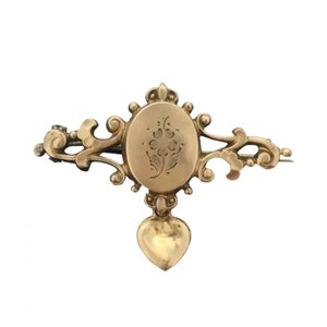 May include: A gold-toned brooch with a heart-shaped pendant. The brooch features an oval-shaped locket with a floral design engraved on the front.