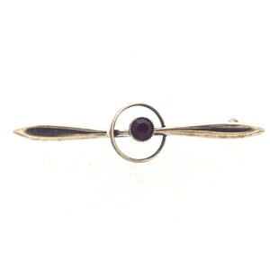 Può includere: Un fermaglio d'oro con una pietra preziosa viola al centro. Il fermaglio ha la forma di un cerchio con due ali sottili e appuntite che si estendono verso l'esterno.