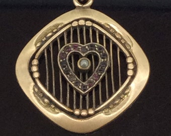 Puede incluir: Un colgante de oro con un diseño en forma de corazón engastado con pequeñas piedras preciosas púrpuras y una sola perla en el centro. El corazón está rodeado por un diseño de jaula con barras verticales.