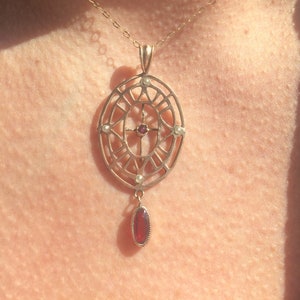 Könnte beinhalten: Eine goldene Halskette mit einem filigranen, kunstvollen Anhänger, der ein Kreuz und kleine Perlen zeigt. Der Anhänger ist oval geformt und hängt an einer dünnen goldenen Kette. Ein einzelner roter Edelstein hängt an der Unterseite des Anhängers.