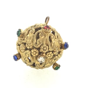 Puede incluir: Un colgante de color dorado con un diseño redondo y ornamentado. El colgante está decorado con cinco piedras de colores: una roja, dos verdes y dos azules.