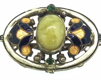 May include: An oval-shaped brooch with a yellow stone surrounded by a silver frame. The frame is decorated with blue and orange enamel and green gemstones.