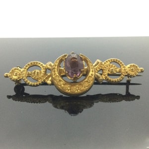 Könnte beinhalten: Eine goldene Halbmond-Brosche mit einem violetten Edelstein in der Mitte. Die Brosche ist mit filigranen Details verziert und hat eine Nadel auf der Rückseite.