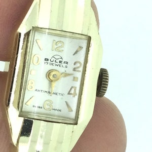 Könnte beinhalten: Eine goldfarbene Armbanduhr mit weißem Zifferblatt und schwarzen Zeigern. Auf dem Zifferblatt der Uhr steht "BULER 17 JEWELS" und "ANTIMAGNETIC".