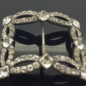 May include: A silver buckle with a square shape and a decorative design of small, clear stones. The buckle has a metal pin that goes through the center.