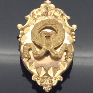 May include: A gold-toned oval-shaped brooch with a detailed ornate design. The center features a wreath with a bow and a textured background.