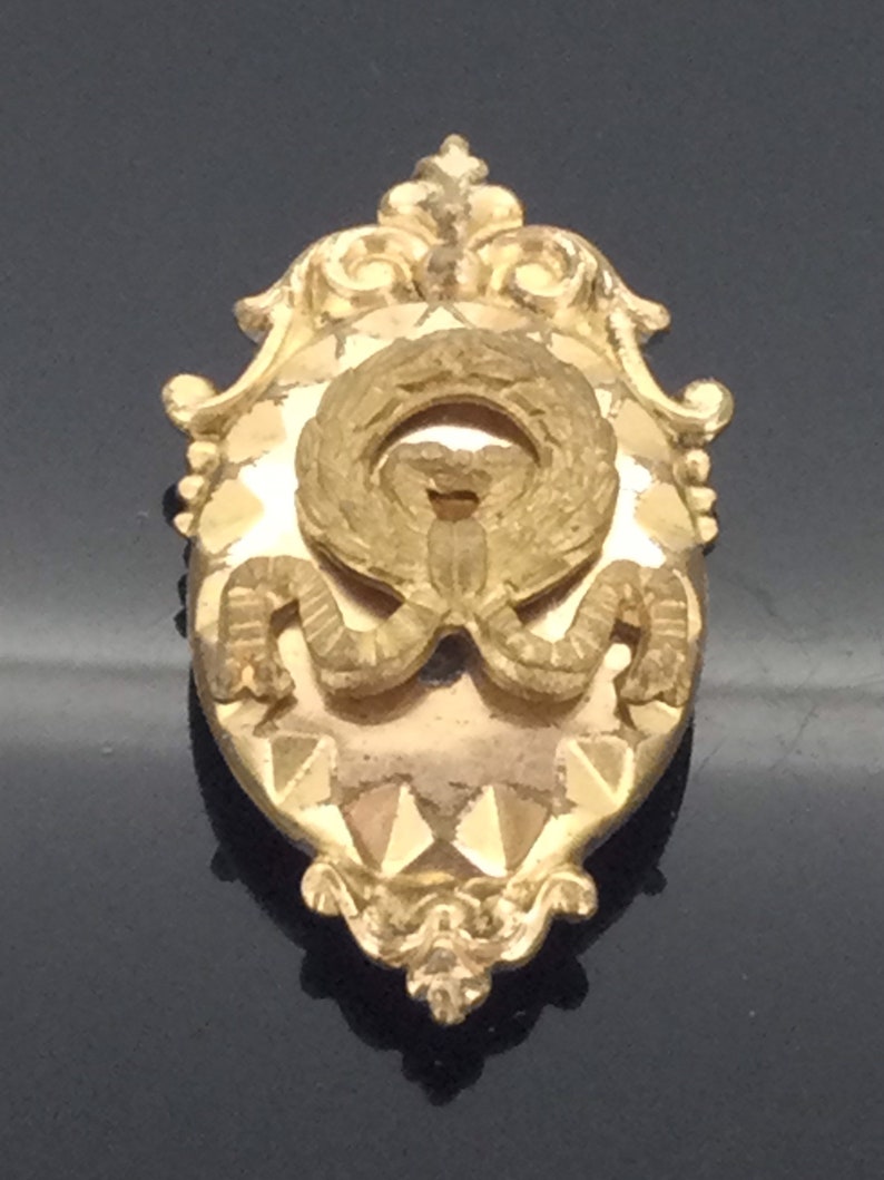 May include: A gold-toned oval-shaped brooch with a detailed ornate design. The center features a wreath with a bow and a textured background.