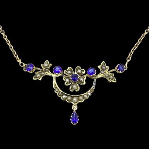 Puede incluir: Un delicado collar de oro con un diseño floral adornado con piedras preciosas azules y perlas. El collar tiene un colgante en forma de lágrima con una piedra preciosa azul.