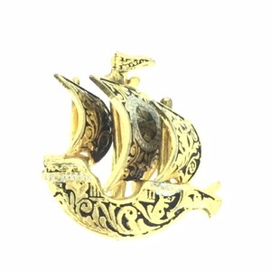 May include: A gold-colored ship brooch with black enamel detailing. The ship has a single sail and intricate floral designs.
