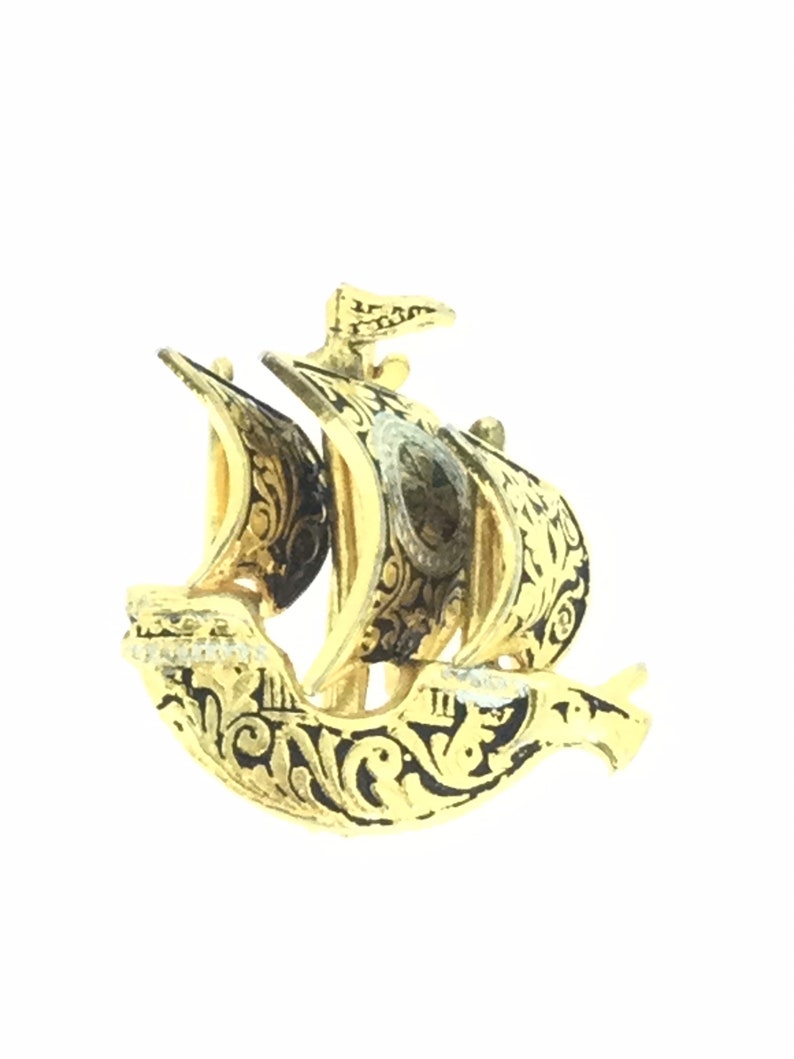 May include: A gold-colored ship brooch with black enamel detailing. The ship has a single sail and intricate floral designs.